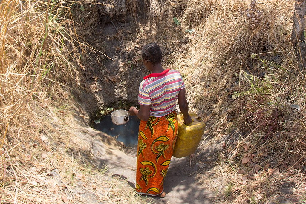 A Zambian woman carries a large, empty, yellow jerry can in her right hand, and a small kitchen pot with a handle in her left. She walks down a rugged path towards a contaminated water hole, a vital but precarious water source for her community.