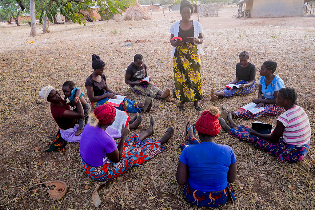 Eight Zambian women from a small village sit outdoors, arranged in a circle on the ground, each holding a Bible on their laps. One woman stands among them, holding a Bible and leading a Bible study session.