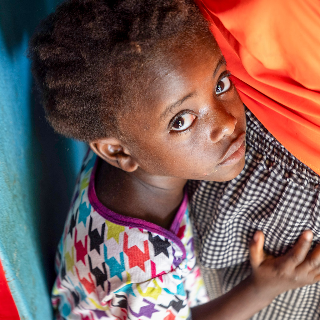 Five-year-old girl, standing close to her mother for comfort, gazes shyly at the camera. Her family, Haitian immigrants, live and work in a Dominican batey.