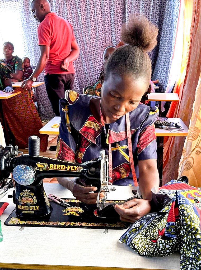 A Zambian woman uses a manual sewing machine as part of the sewing project group. other women and the project leader can be seen in the background.